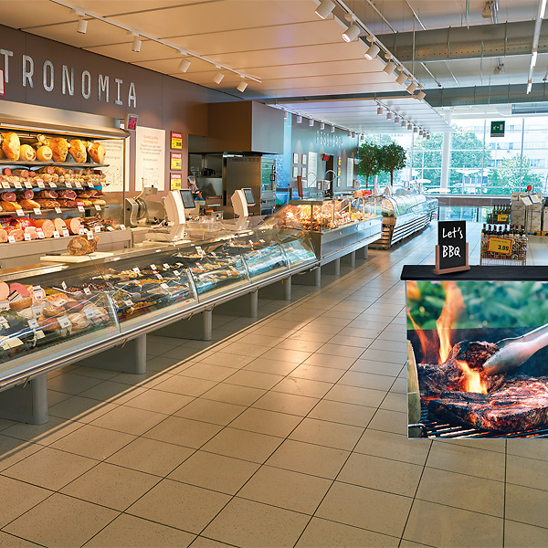 Fødevaredisplay i supermarked med kød, ost og grilltema foran.
